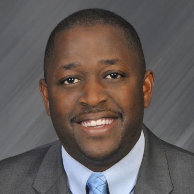 A smiling darker-skinned individual with no hair wearing a gray suit and tie against a gray background