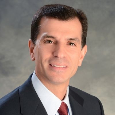 A smiling light-skinned male with dark brown hair wearing a navy suit and tie against a gray marbled background