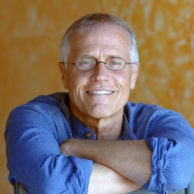 A smiling light-skinned male with white hair wearing glasses and a blue shirt against a yellow background