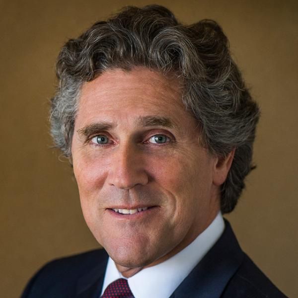 A smiling light-skinned male with gray hair and a dark suit against a brown gradient background