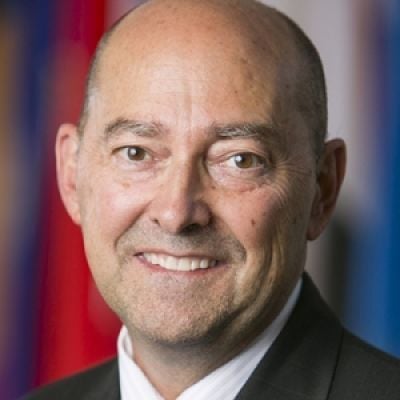 A smiling light-skinned male with no hair wearing a dark suit jacket against an abstract background