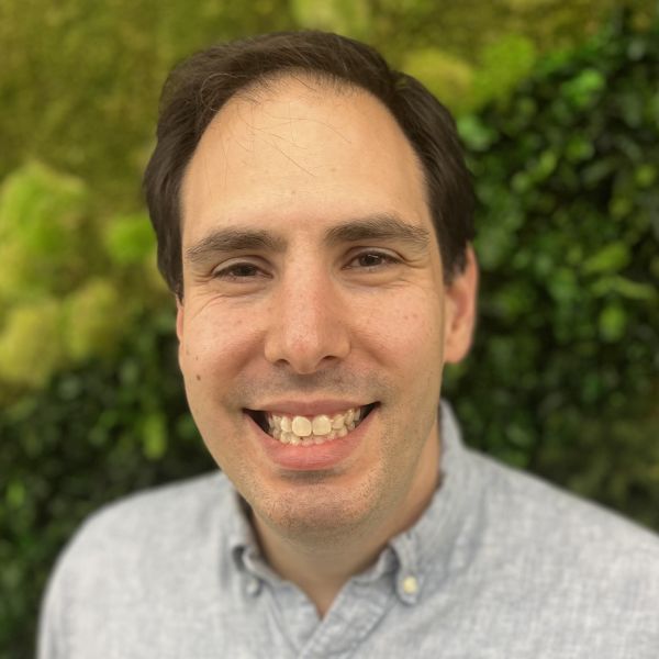 Smiling light-skinned, male-presenting individual with short dark hair wearing a grey shirt on an outdoor backdrop.