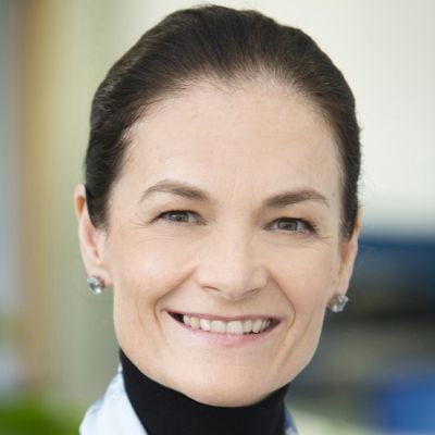 A smiling light-skinned female with dark brown hair wearing a black turtleneck against a blurred background