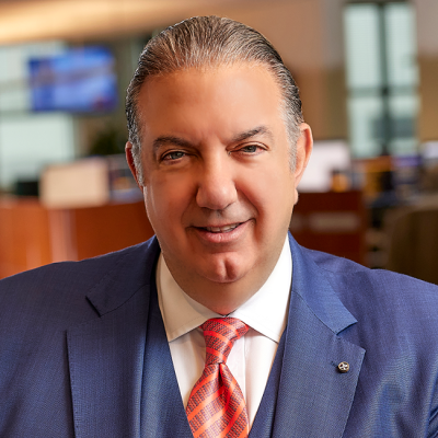 A smiling light-skinned male with gray hair wearing a three-piece blue suit against a blurred background