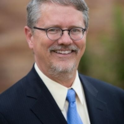 A smiling light-skinned male with gray hair and a goatee wearing a dark blue suit and tie against an outdoor setting