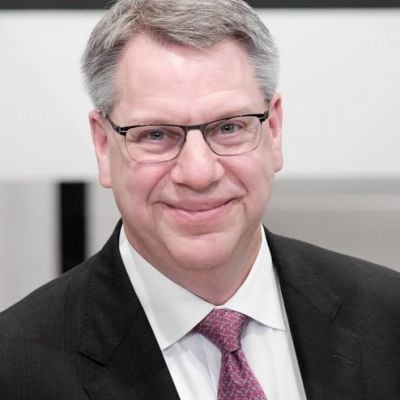 A smiling light-skinned male with gray hair wearing glasses and a black suit and tie against a blurred background