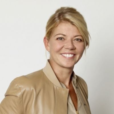 A smiling light-skinned female with blonde hair wearing a beige leather jacket against a cream background