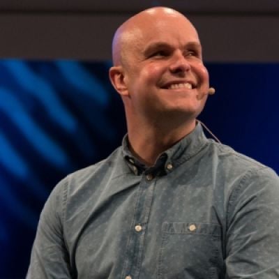 A smiling light-skinned male with a microphone wearing a gray shirt against a dark blue background