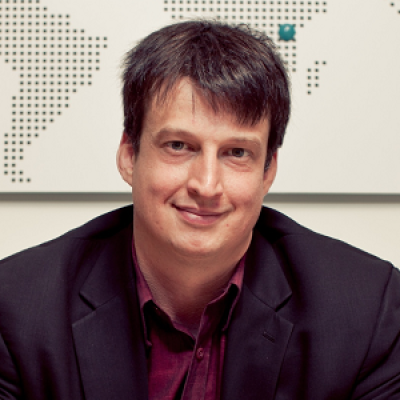 A smiling light-skinned male with brown hair wearing a dark suit jacket against an ambiguous background