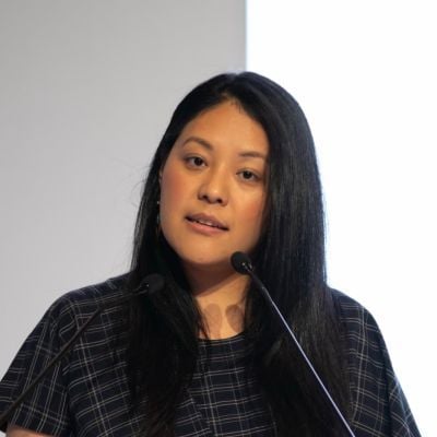 A female with long black hair wearing a black patterned shirt standing a microphone speaking