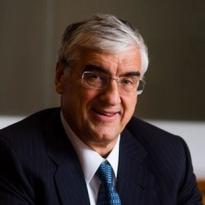 A smiling light-skinned male with gray and white hair wearing a dark suit and tie against an ambiguous background