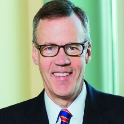 A smiling light-skinned male with gray hair wearing glasses and a black suit and tie against a green background