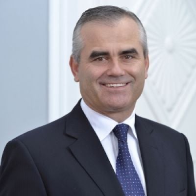 A smiling light-skinned male with gray hair wearing a dark suit and tie against a gray background