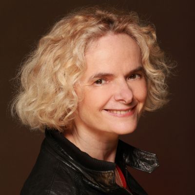 A smiling light-skinned female with short blonde curly hair wearing a black leather jacket against a dark brown backdrop