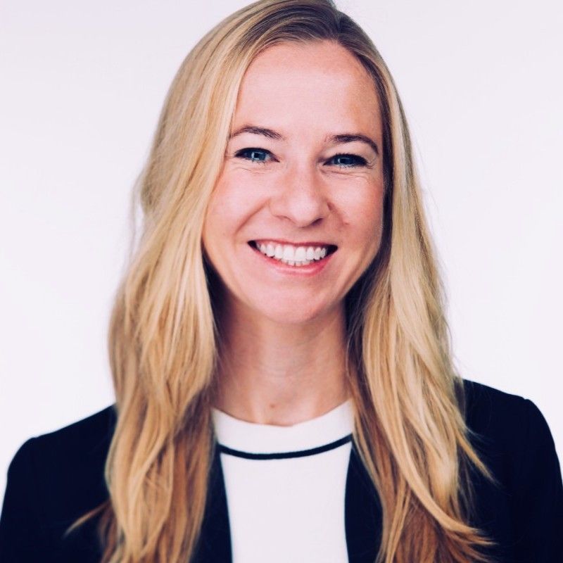 A smiling light-skinned female with long blonde hair wearing a white blouse and a black jacket against a light background