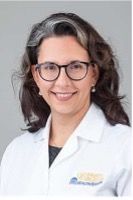 A smiling female with light skin and dark gray and brown hair wearing glasses and a lab coat against a light gray background