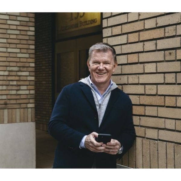 A smiling light-skinned male with light hair wearing a navy blue jacket over a gray sweater leaning against a brick wall holding his cell phone