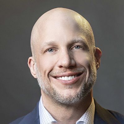 A smiling light-skinned male with no hair and a short gray beard wearing a navy suit jacket against a dark gray background