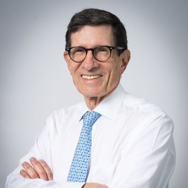 A smiling light-skinned male with dark brown hair wearing glasses and a white button-up shirt with a blue tie against a white gradient background