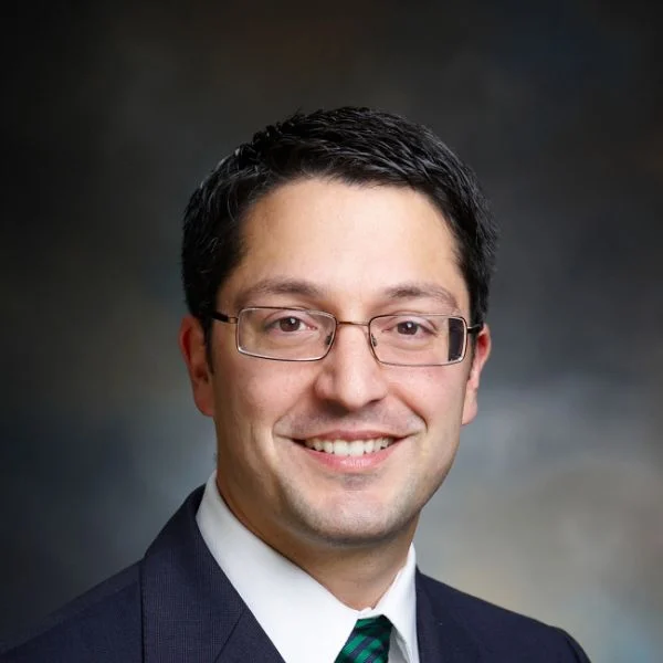 A smiling light-skinned male wearing a dark brown hair wearing glasses and a dark suit jacket and tie against a gray gradient background