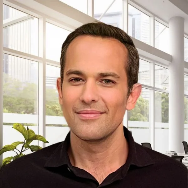 A smiling light-skinned male with dark brown hair wearing a black shirt against a set of windows in the background