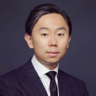 A light-skinned male of Asian heritage with short black hair wearing a dark suit and tie against a dark gray background