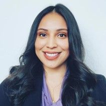 A smiling female with long black hair wearing a purple shirt and black jacket against a gray background