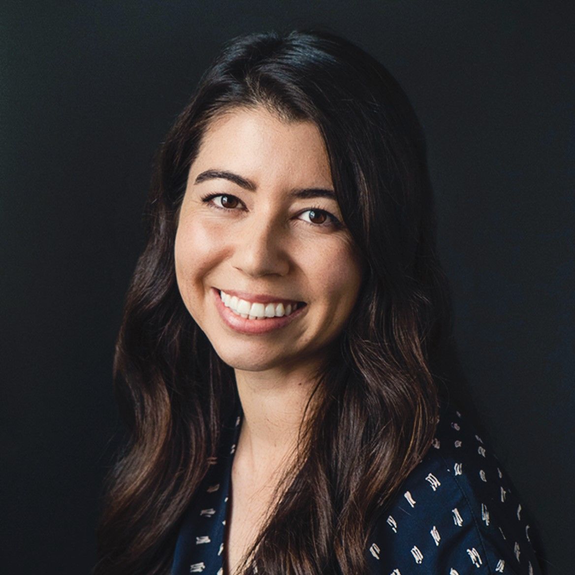 A smiling light-skinned female with long dark brown hair wearing a navy shirt against a gray background