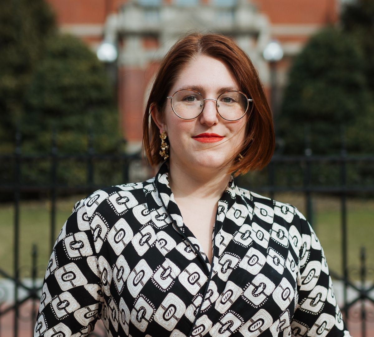 An woman with auburn hair and glasses in front of a building. 