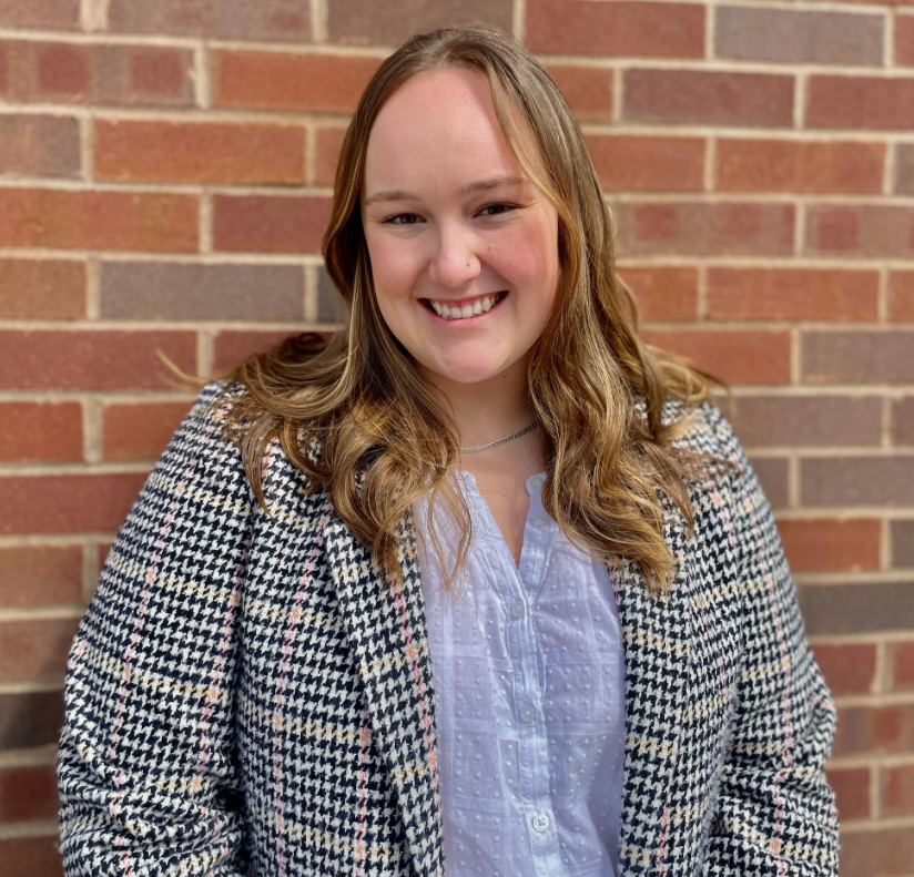 A woman with light hair and a blazer against a brick background.