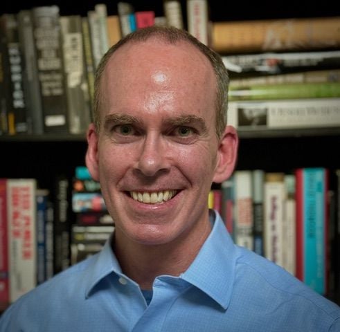 A smiling man in a blue blouse against a backdrop of books.