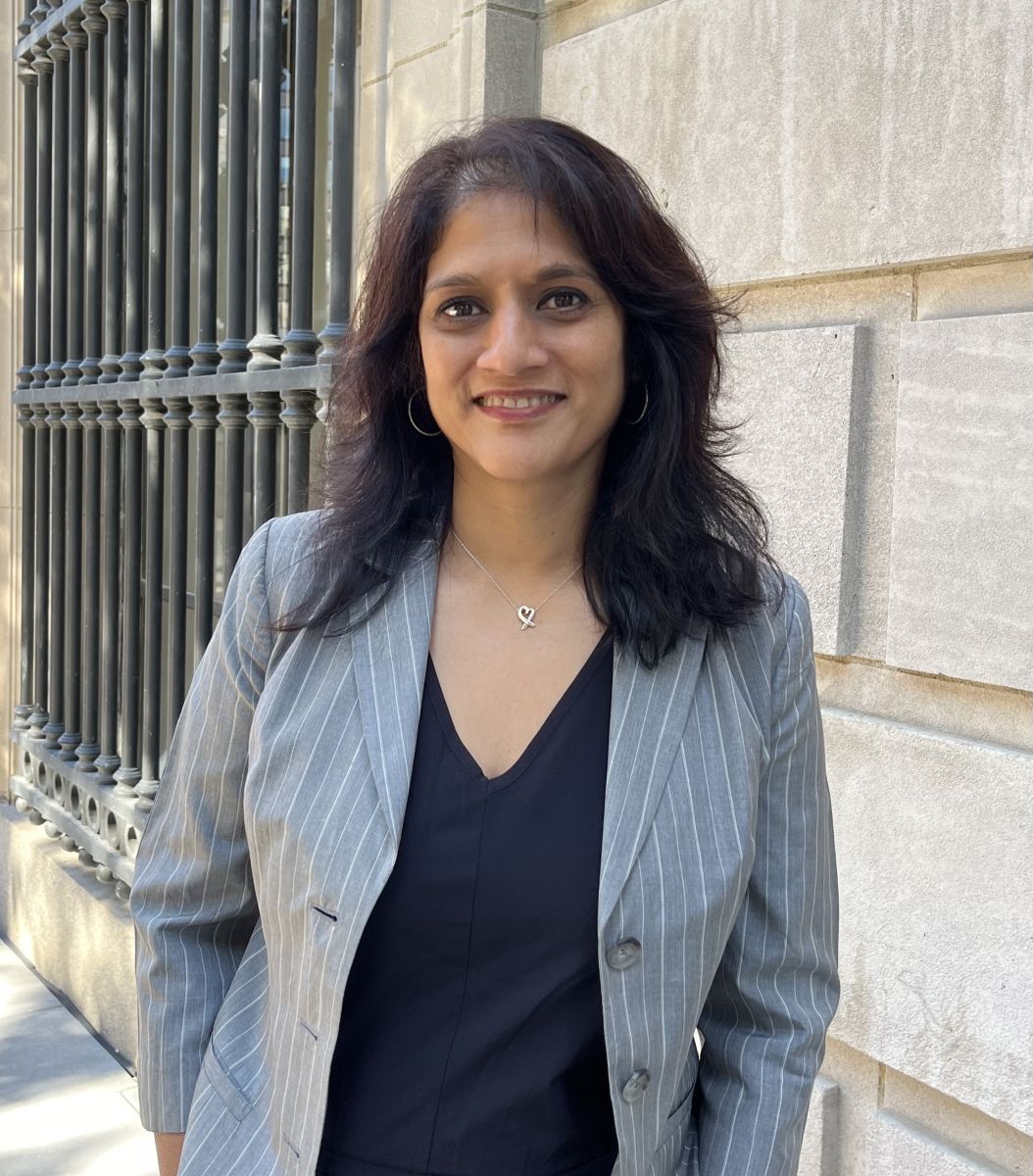 A smiling darker-skinned female with medium length black hair wearing a gray blazer over a black shirt against an ambiguous background