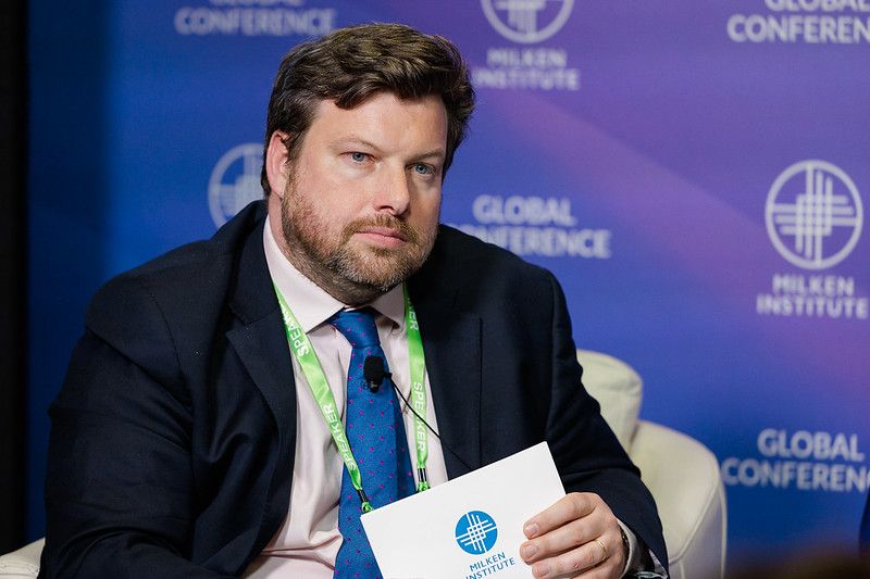 A male conference presenter against the the Milken Institute Global Conference backdrop.