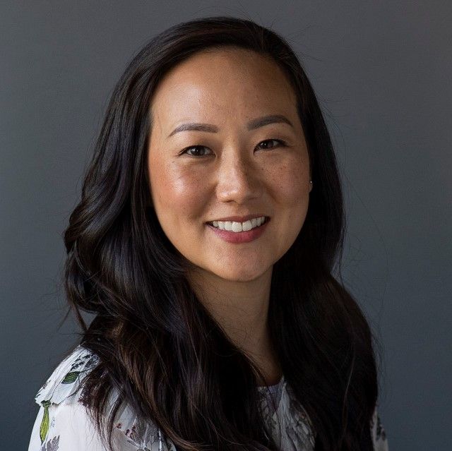 A smiling woman of Asian heritage with long black hair wearing a white shirt against a gray background