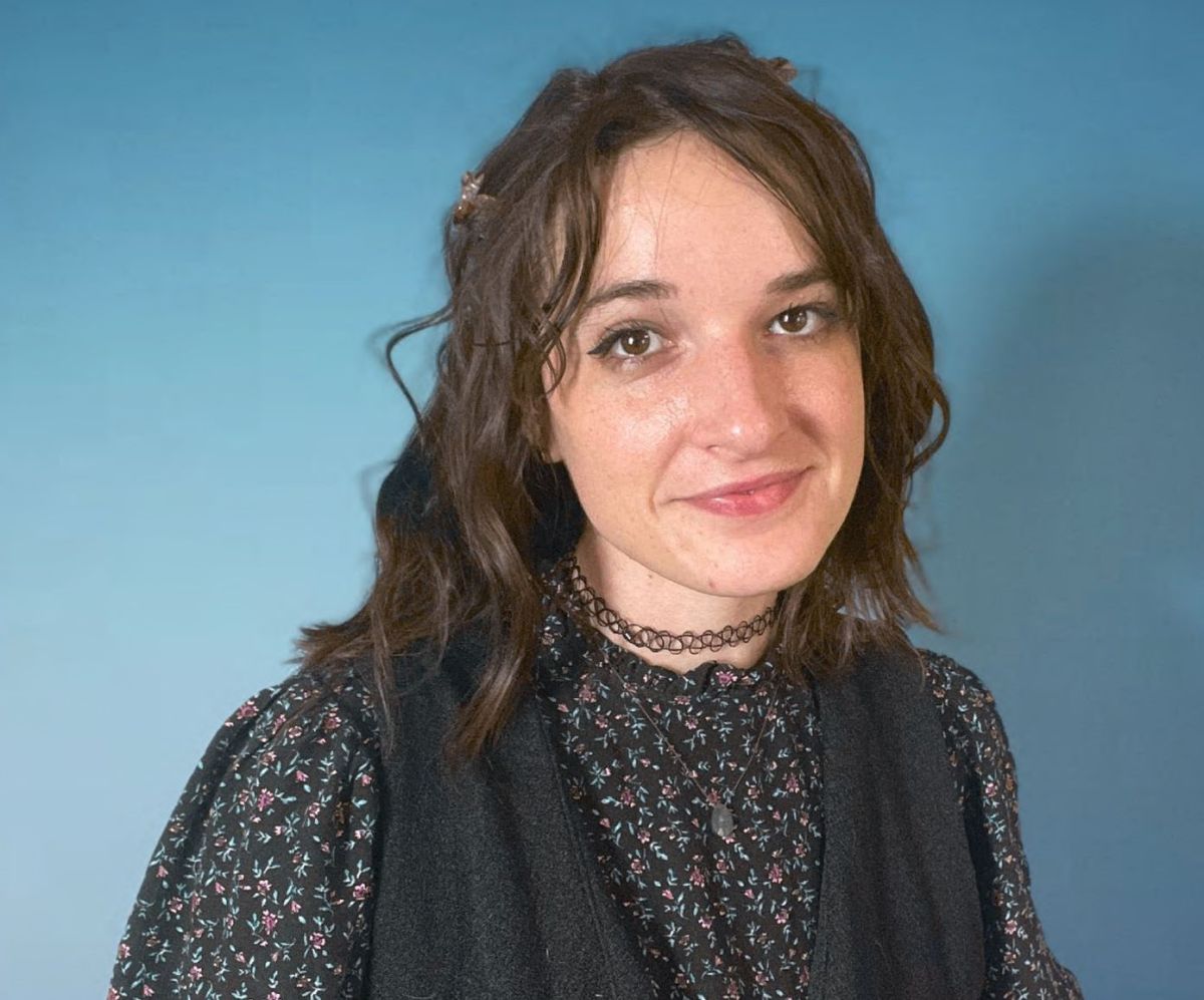 A light-skinned female with brown hair wearing a dark outfit against a blue background.