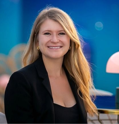 person with long blonde hair and a black blazer in front of blue backdrop