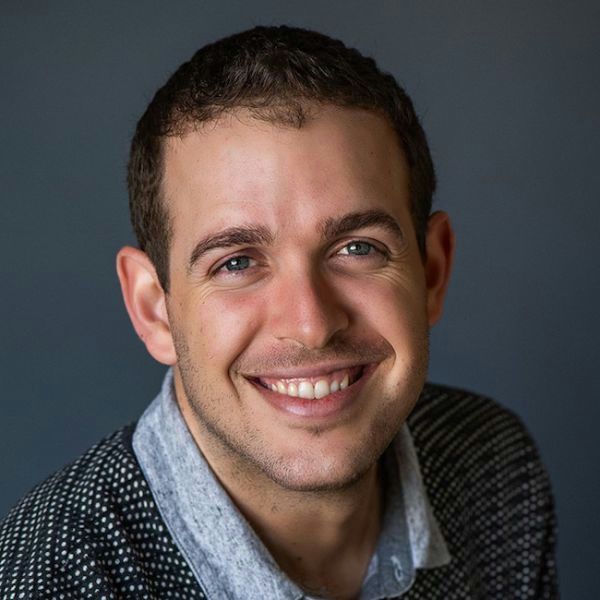 Person with close cut short hair leaning into camera, smiling, wearing knit sweater over blue shirt