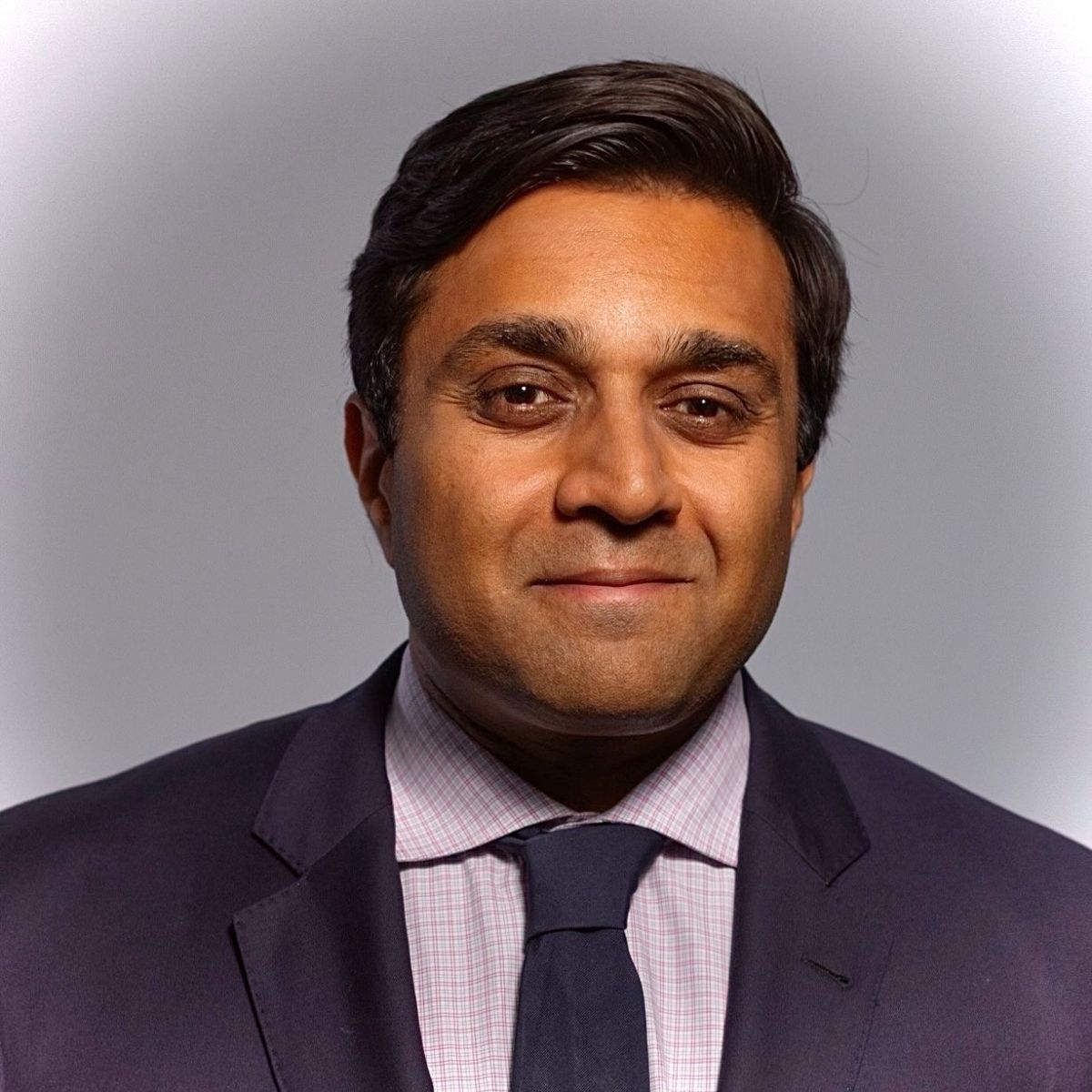 A headshot of an adult man with dark skin who is possibly Indian in appearance. He has combed black hair, great eyebrows, and he is slightly smiling at the camera. He is wearing a dark suit with a dark blue tie. 