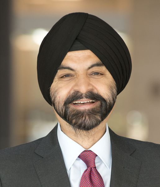 A headshot of a smiling adult man with white skin, a full black beard, and wearing a black pagri. He is also wearing a black suit with a white collared shirt and a red tie.