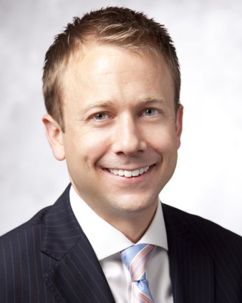 A headshot of an adult man with white skin and short brown-blonde hair. He is smiling at the camera and wearing a dark suit with a collared shirt and a pink tie. 