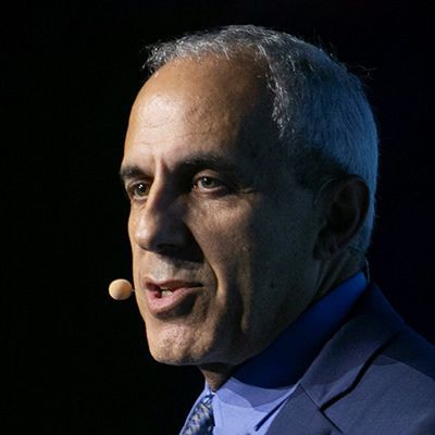 An image of an older adult man with white and slightly tan skin and short white hair. It is an image of mostly his head and he is speaking into an earpiece microphone and wearing a blue suit and tie. He is against a black background. 