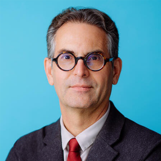 A headshot of an older adult man with short gray hair and white skin. He is wearing circular glasses, a gray suit, and a red tie.