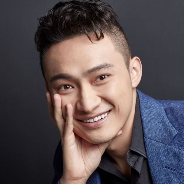 A headshot of a smiling adult man who is Asian in appearance. He has black hair with a fade, brown eyes, and is leaning on his hand. 