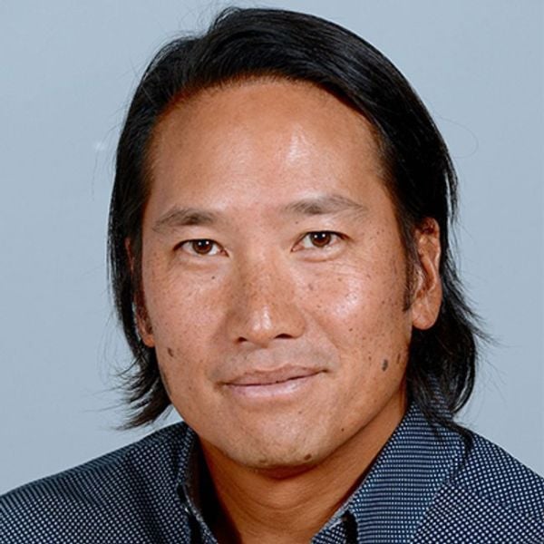 A headshot of an adult man who is Asian in appearance, with long-ish black hair and a blue collared shirt. He is slightly smiling at the camera.