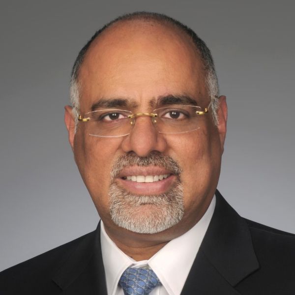 An image of an adult man with dark skin who has grayish hair on the sides of his head and a grayish goatee. He is smiling at the camera and wearing a suit with a blue tie as well as rectangular clear glasses.