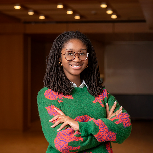 A smiling dark-skinned woman named Adoma Addo with shoulder-length dark hair, wearing glasses as well as a green sweater with pink tigers, and standing in a room with dark wood details.