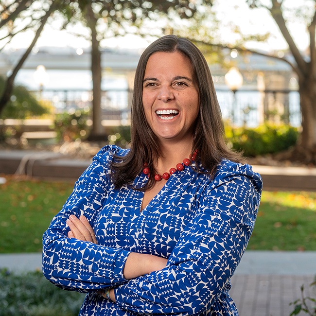 A smiling light-skinned female named Cara Altimus with shoulder length dark brown hair wearing a vibrant blue shirt and red necklace, standing with her arms crossed against an ambiguous outdoor background.