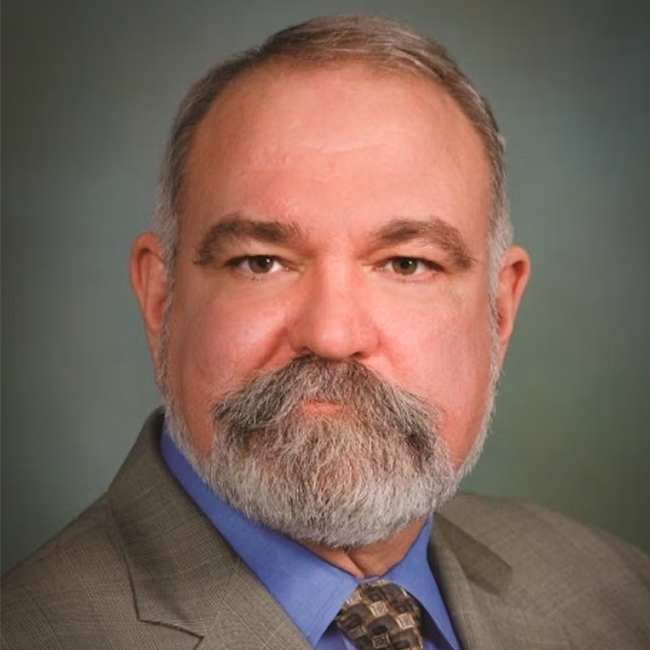 A photo of an older adult man with light white skin and gray hair, including a gray moustache and beard. He is wearing a suit and has brown-ish eyes. 