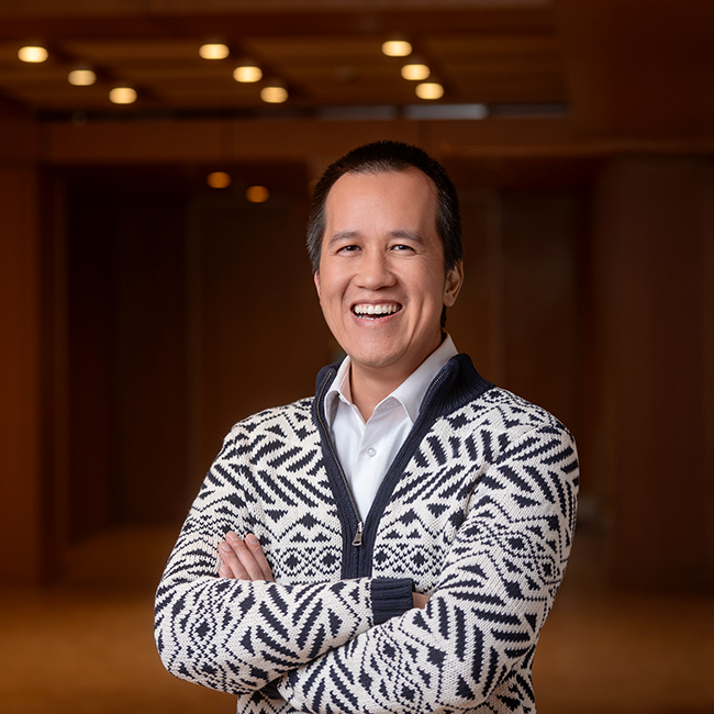 A smiling light-skinned, Asian-presenting man named Daniel Pham with short black hair, wearing a geometric black and white sweater, and smiling and standing with his arms crossed in a room with dark wood details.