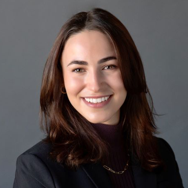 An image of a smiling adult woman named Elana Jacob, who is an  associate director on the Business and Program Development team at the Milken Institute. She has shoulder-length brunette hair, light white skin, and is wearing a black shirt against a gray background. 